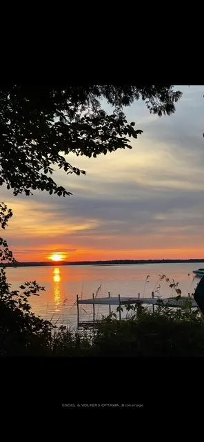 Sunset over a calm lake, framed by trees. A small dock extends into the water. The sky is a mix of orange, pink, and gray.