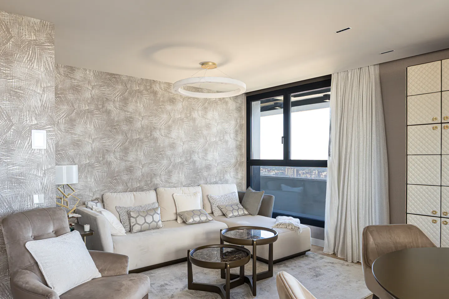 A modern living room with a beige sectional sofa, round tables, and a large window with city views. The walls have a textured wallpaper.