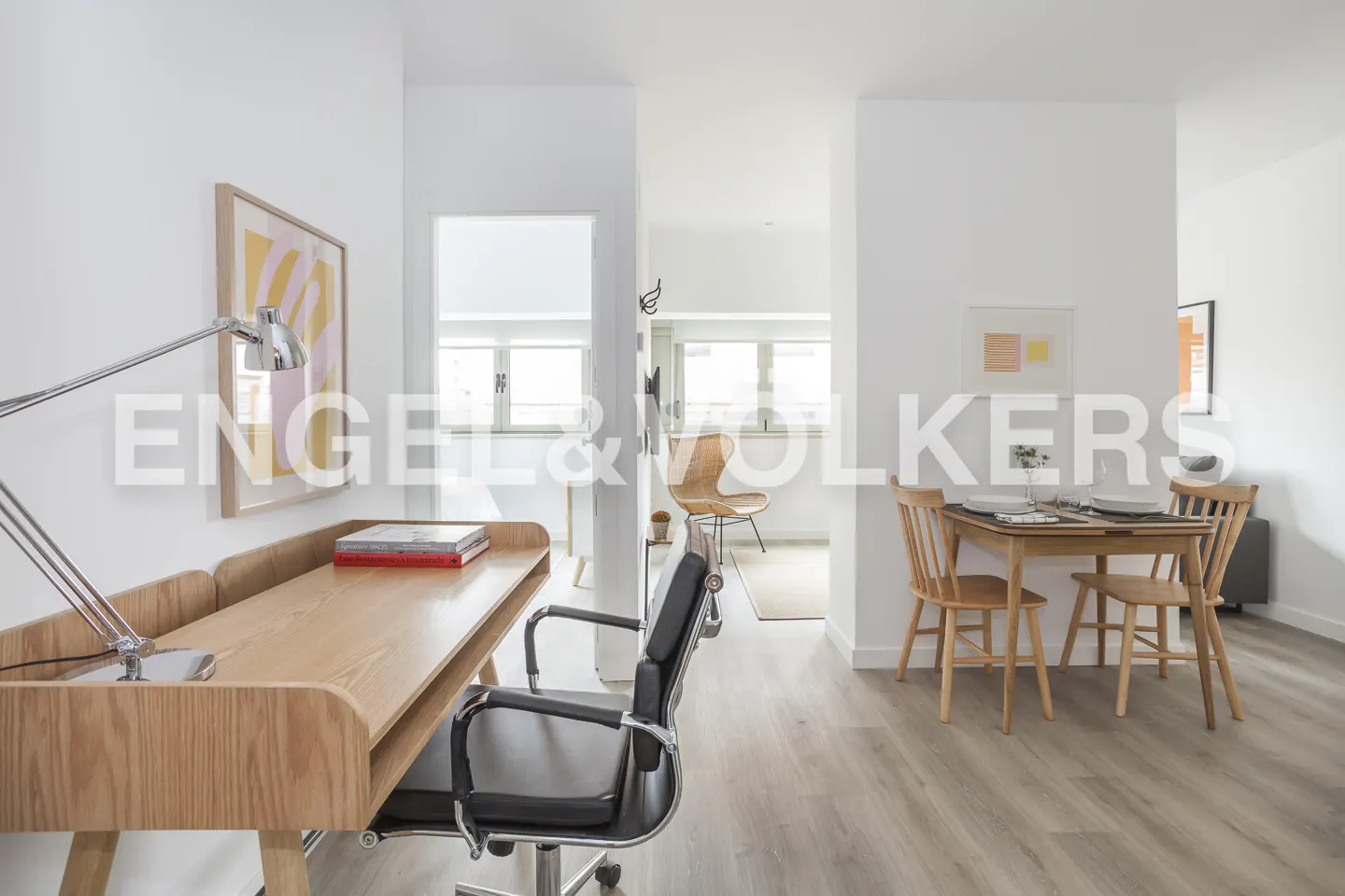 Bright apartment interior with a wooden desk, black chair, and a dining table set for two, all on light wood-look flooring.