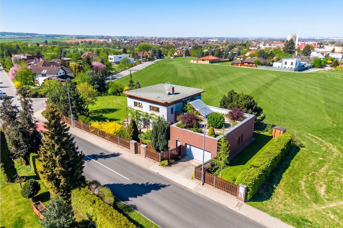 Aerial view of a modern two-story house with a green roof and solar panels, surrounded by a wooden fence and lush greenery.
