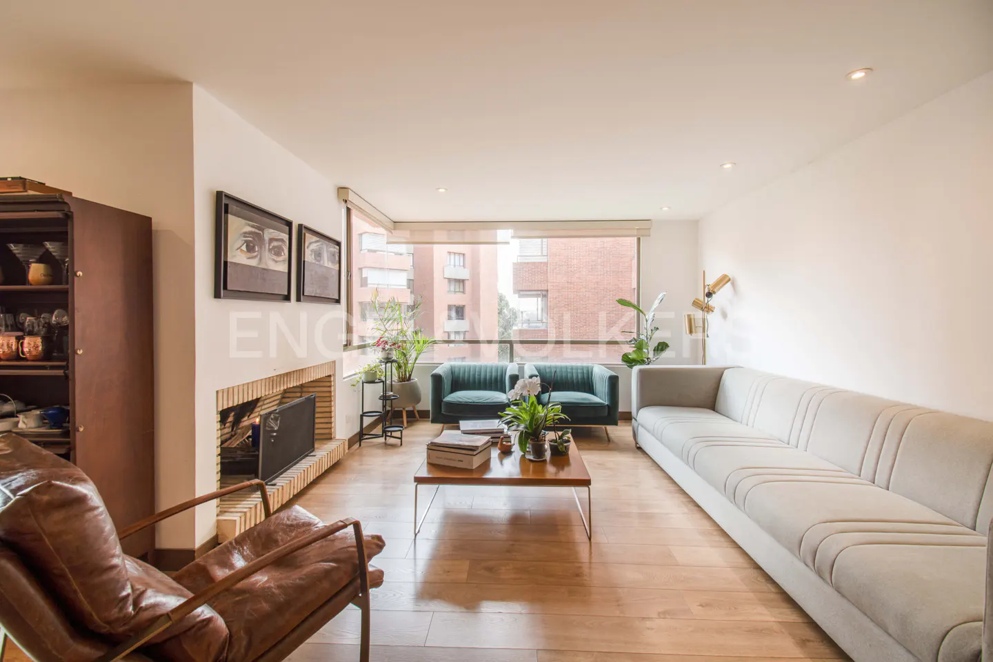 Bright living room with hardwood floors, a gray sofa, two green loveseats, and a brown leather chair. A fireplace and large windows are visible.
