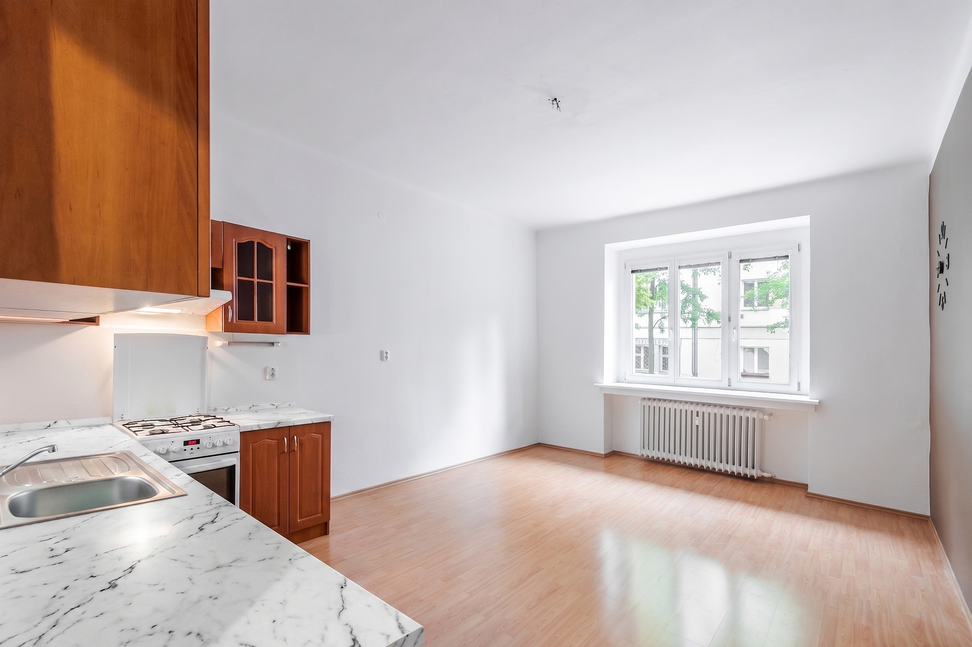 Bright kitchen with wood cabinets, marble-look countertops, white walls, light wood floors, and a window with a radiator.