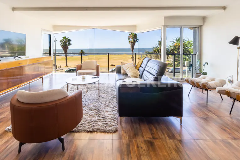 Bright living room with ocean view. Black leather sofa, brown chairs, and wood floors. Palm trees visible through large windows.
