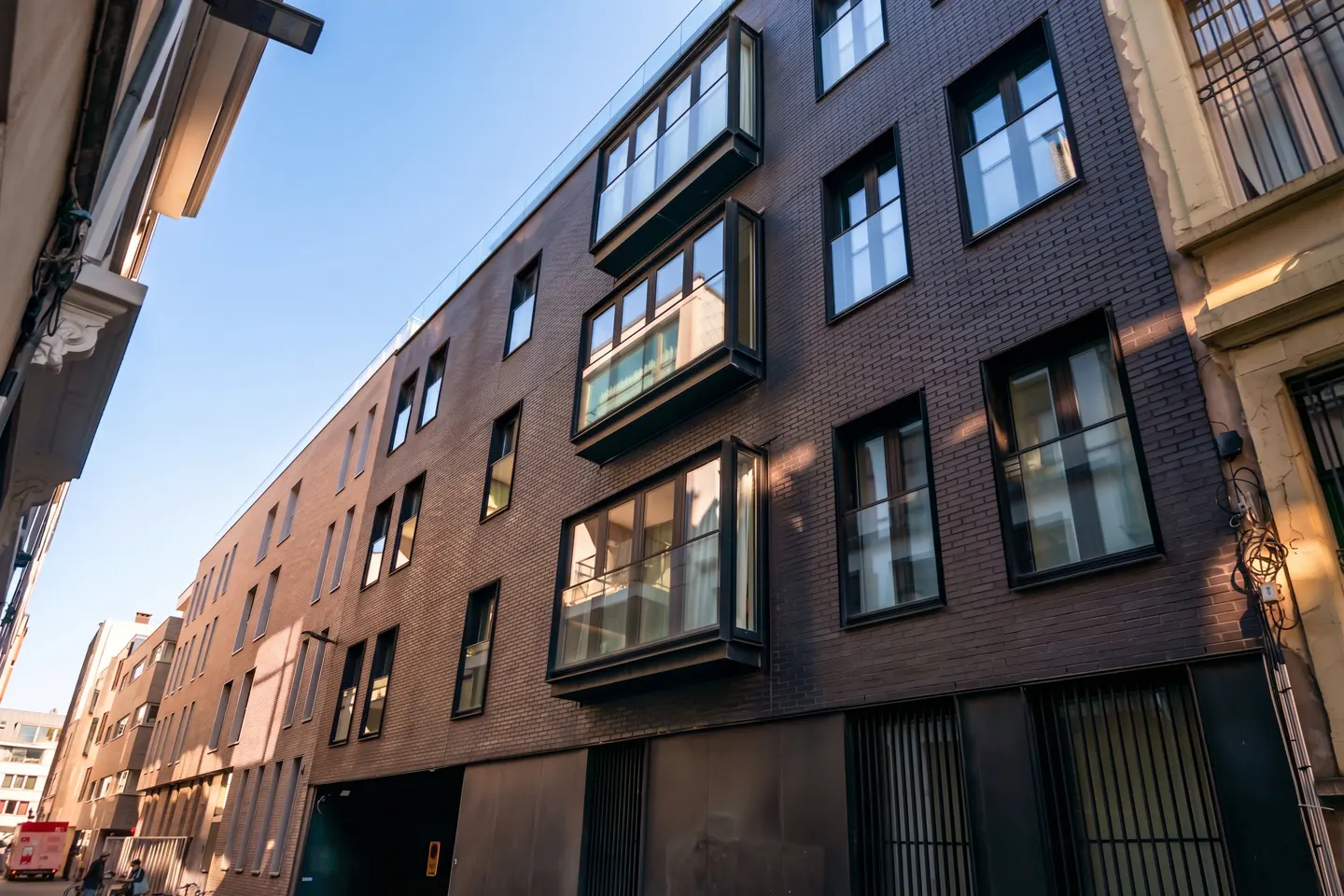 Exterior view of a modern, multi-story brown brick building with black framed windows and glass balconies on a sunny day.