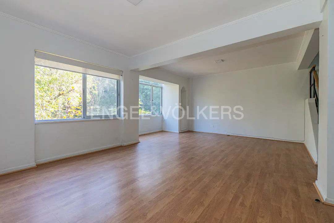 Bright, empty room with wood floors and white walls. Large windows show trees outside. Engel & Völkers logo is visible.