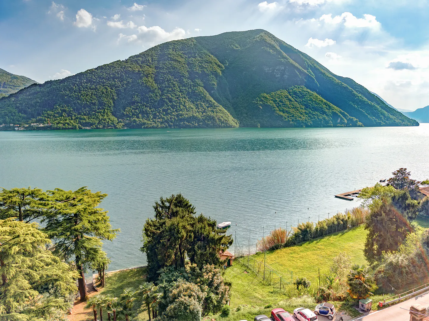 Lake Lugano view with green mountain backdrop. Trees, grass, and parked cars in the foreground. Blue sky with clouds.