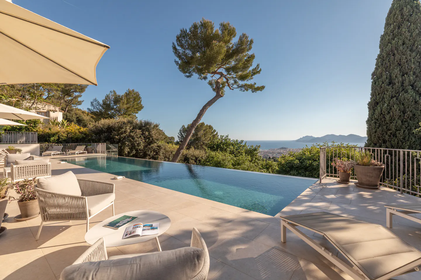 Outdoor patio with infinity pool overlooking the ocean. Lounge chairs and seating area with table and magazines. Trees and blue sky in the background.