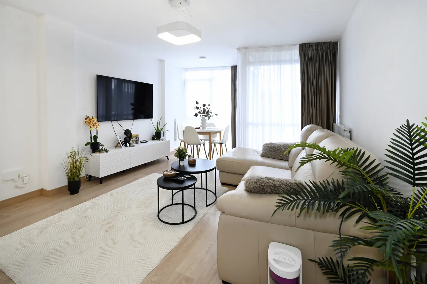 Bright living room with a beige sectional sofa, black coffee tables, and a white media console under a large TV. Dining table in background.
