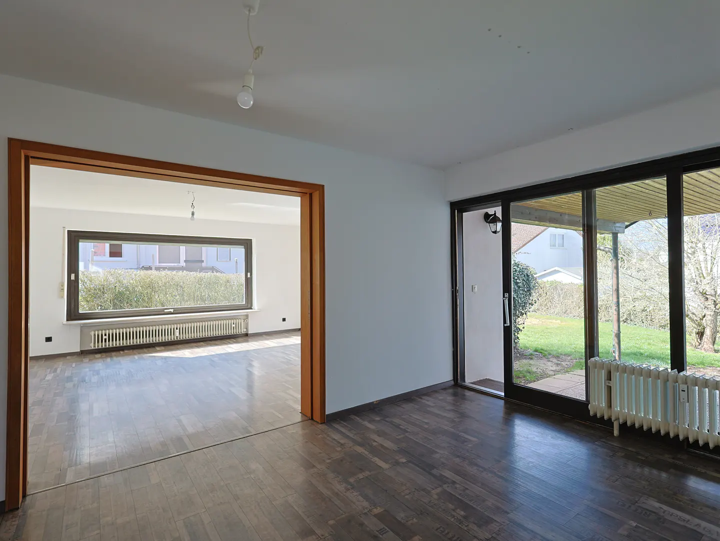 Empty room with dark wood floors, white walls, and a large window. A sliding glass door leads to a patio and garden.