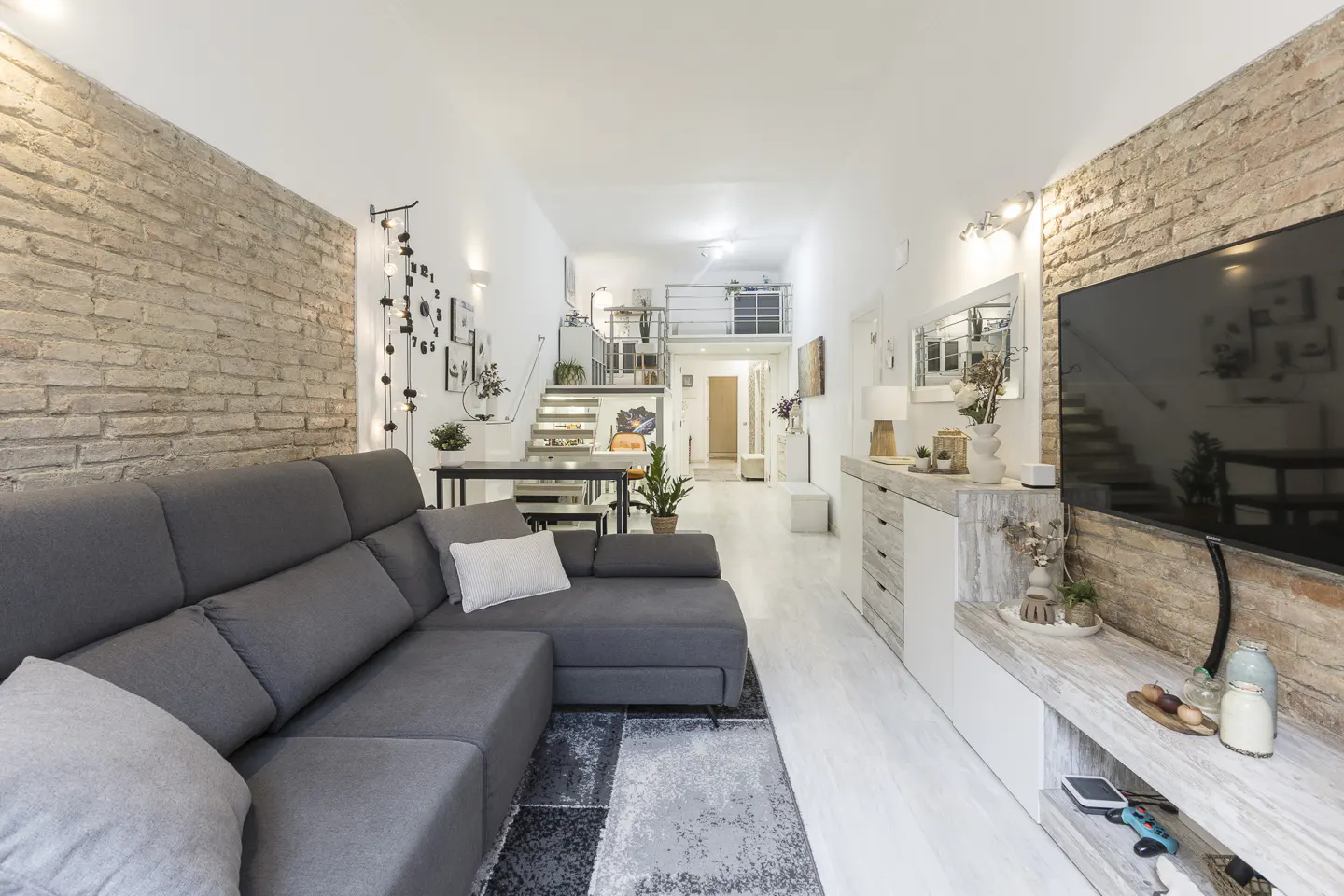 Bright, modern living room with a gray sectional sofa, brick walls, and white wood floors. A staircase leads to a loft area. A large TV is mounted on the wall.