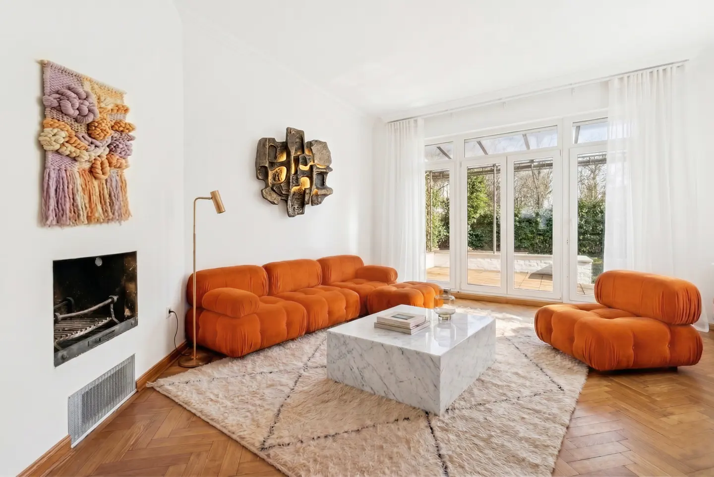 Bright living room with orange sectional sofa, marble coffee table on patterned rug, and white walls with art. French doors lead to a garden.