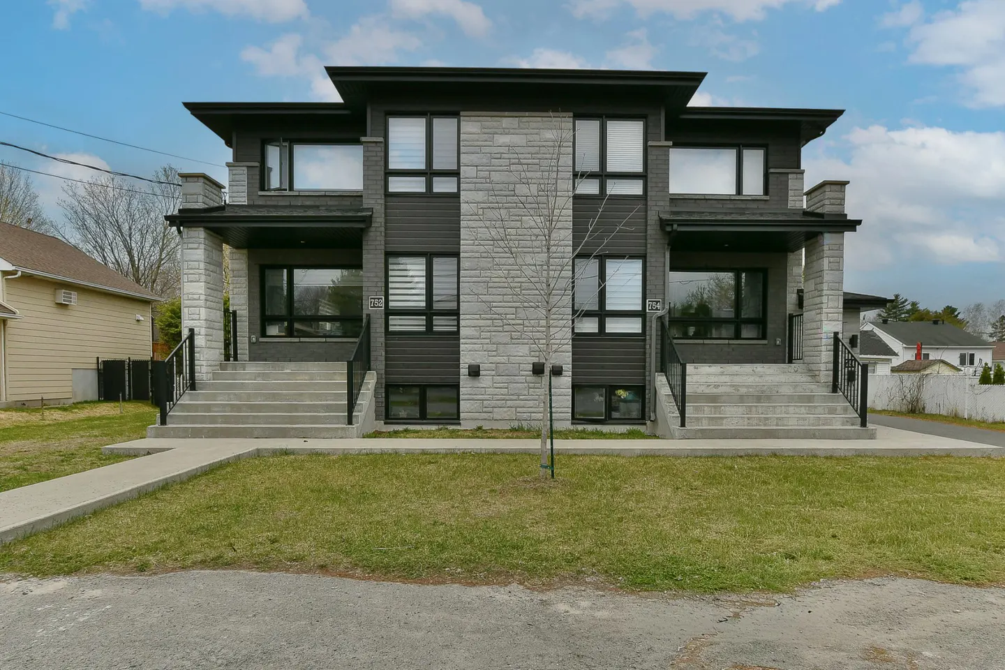 Two-story duplex with gray stone and dark siding, black trim, and concrete steps leading to each entrance. A small tree is centered between the units.