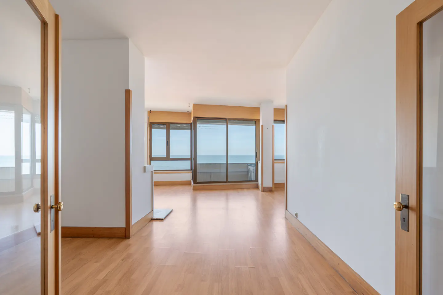 A bright, empty room with wood floors and white walls, seen through open wood-framed doors. Large windows overlook the ocean.