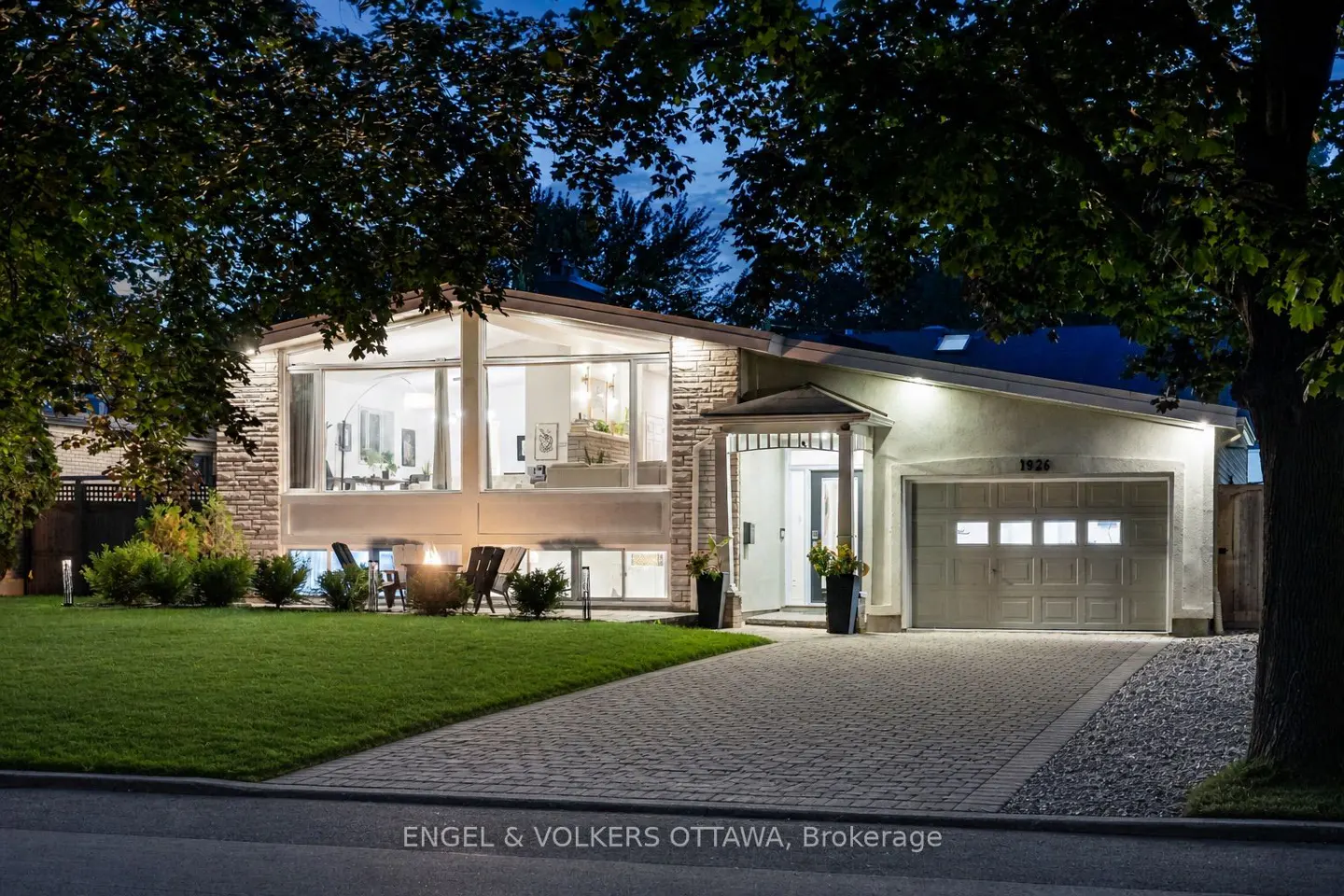 Exterior view of a one-story house with a stone facade and a paved driveway at dusk. A fire pit is on the lawn.