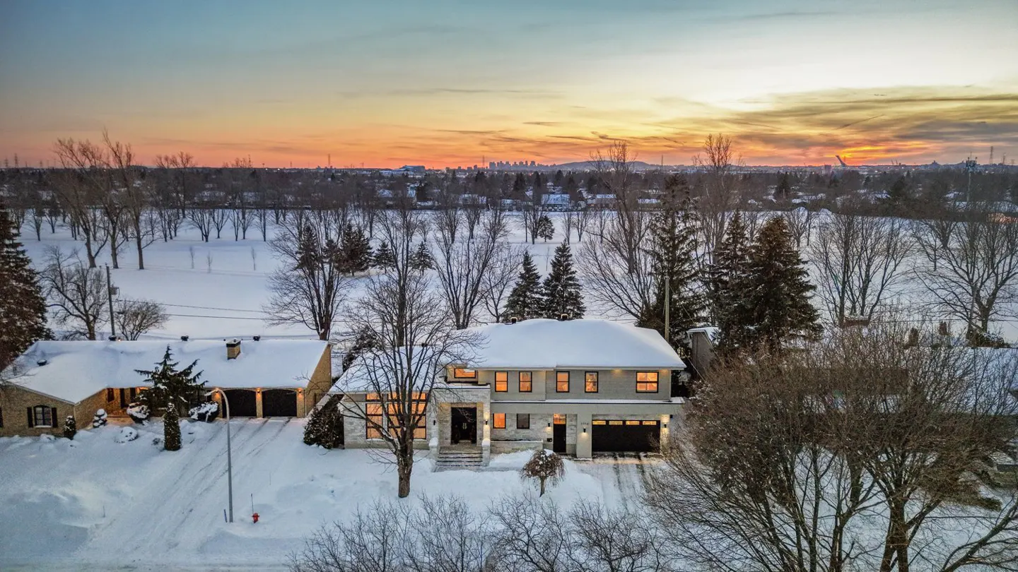 Aerial view of a snow-covered house at sunset. The sky is orange and yellow. Trees surround the house.