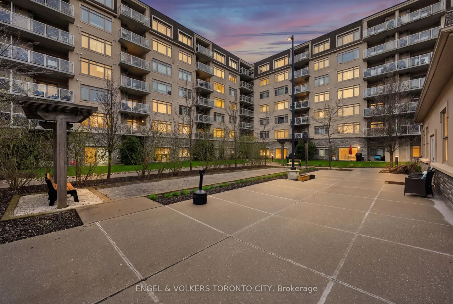 Courtyard of a multi-story apartment building at dusk with a paved patio, benches, and trees under a colorful sky.