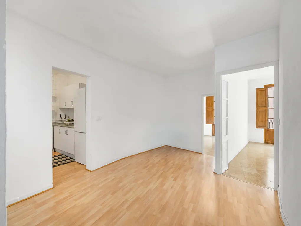 Bright, empty room with light wood floors and white walls. A kitchen and a room with open shutters are visible through doorways.