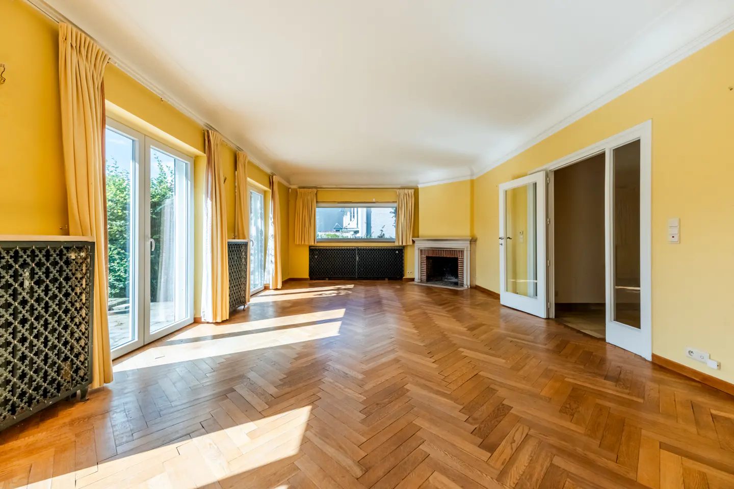 Bright living room with yellow walls, herringbone wood floors, a fireplace, and glass doors leading to a garden.