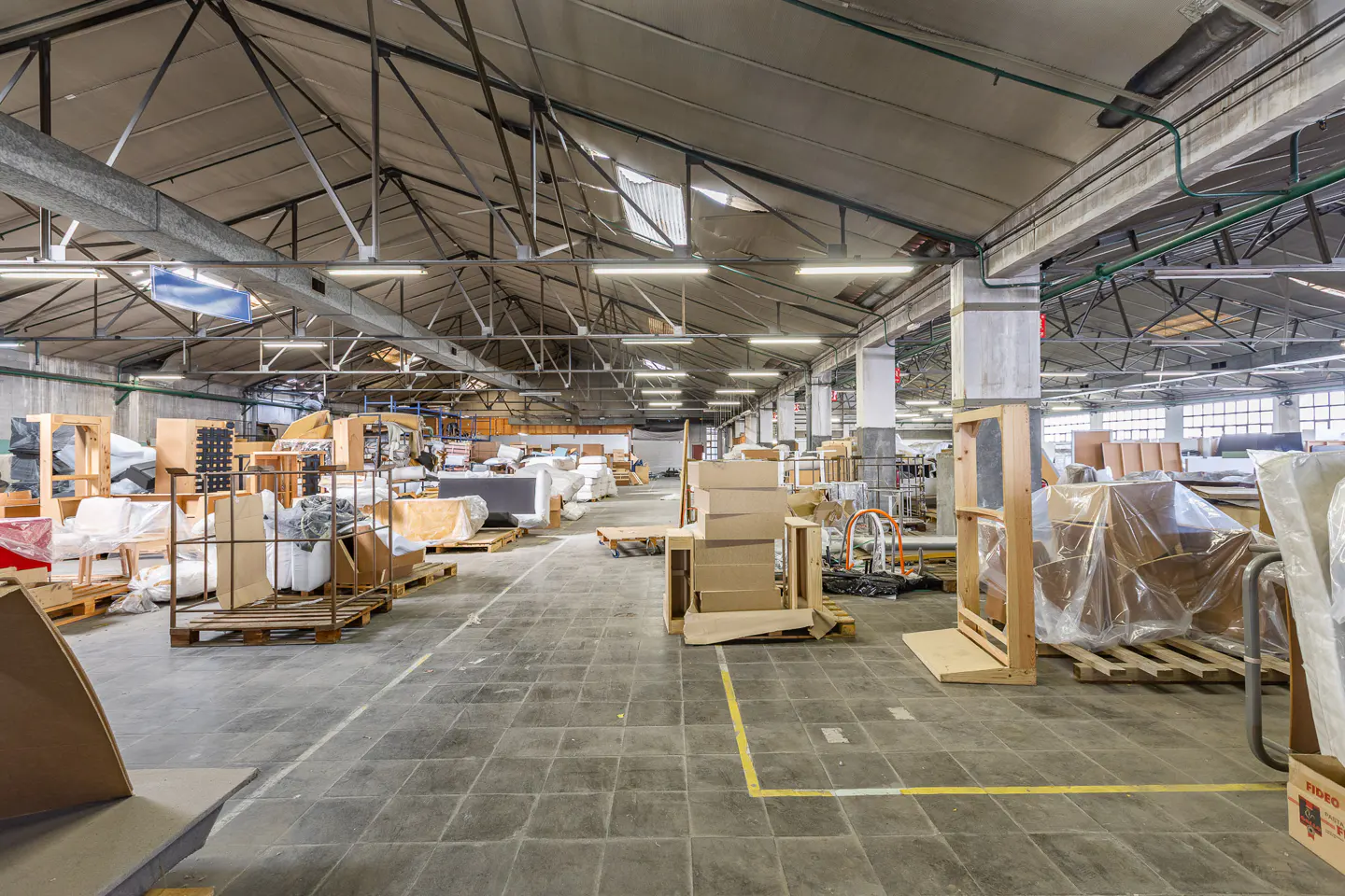 Large industrial warehouse interior with high ceilings, concrete floors, and various furniture items stored on wooden pallets.