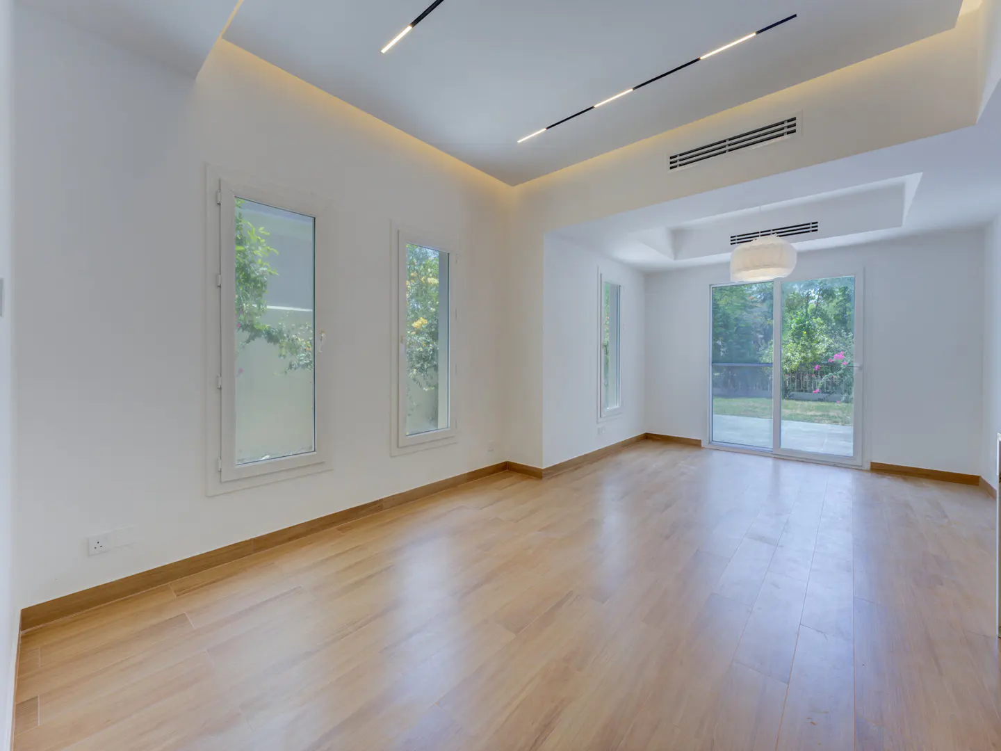 Bright, empty room with wood floors, white walls, and three windows. Sliding glass doors lead to a green yard. Modern lighting fixtures on the ceiling.