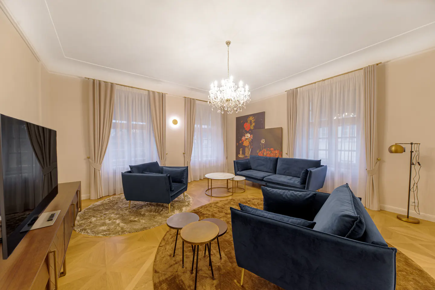 A living room with blue velvet sofas, a chandelier, and beige curtains. There are two paintings on the wall.