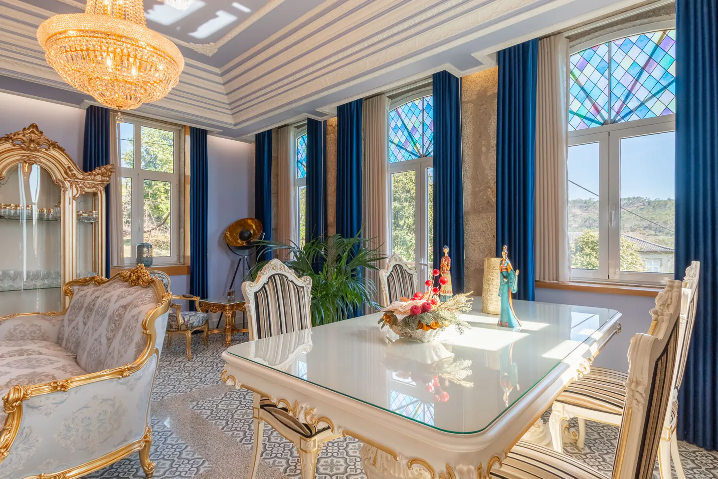 Ornate dining room with a large chandelier, blue curtains, and a white and gold table set for a meal. Stained glass windows add color.