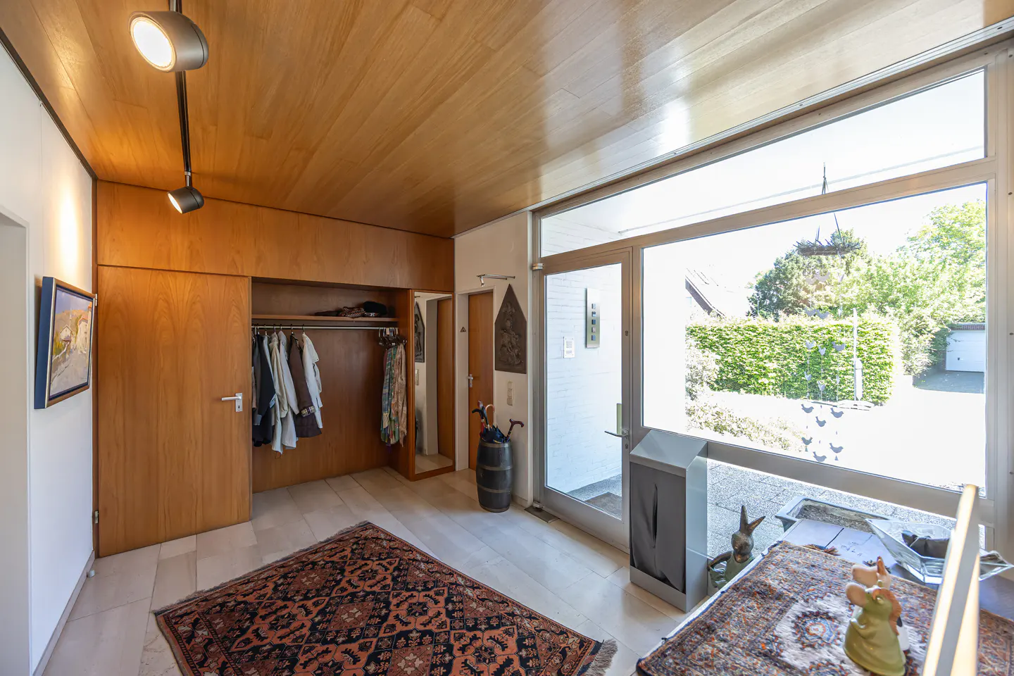 A modern entryway with wood paneling, a large window, and patterned rugs. A closet with clothes is visible.