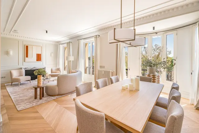 Bright, open-concept living and dining room with light wood floors, neutral furniture, and large windows. A long dining table sits in the foreground.