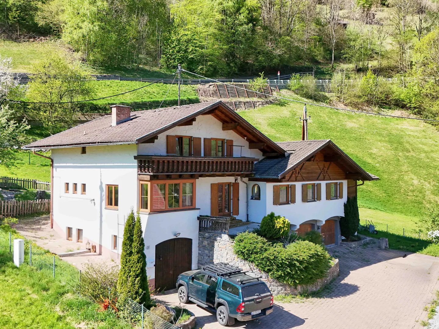 A white two-story house with brown shutters and a balcony, nestled in a green, hilly landscape. A green SUV is parked in front.