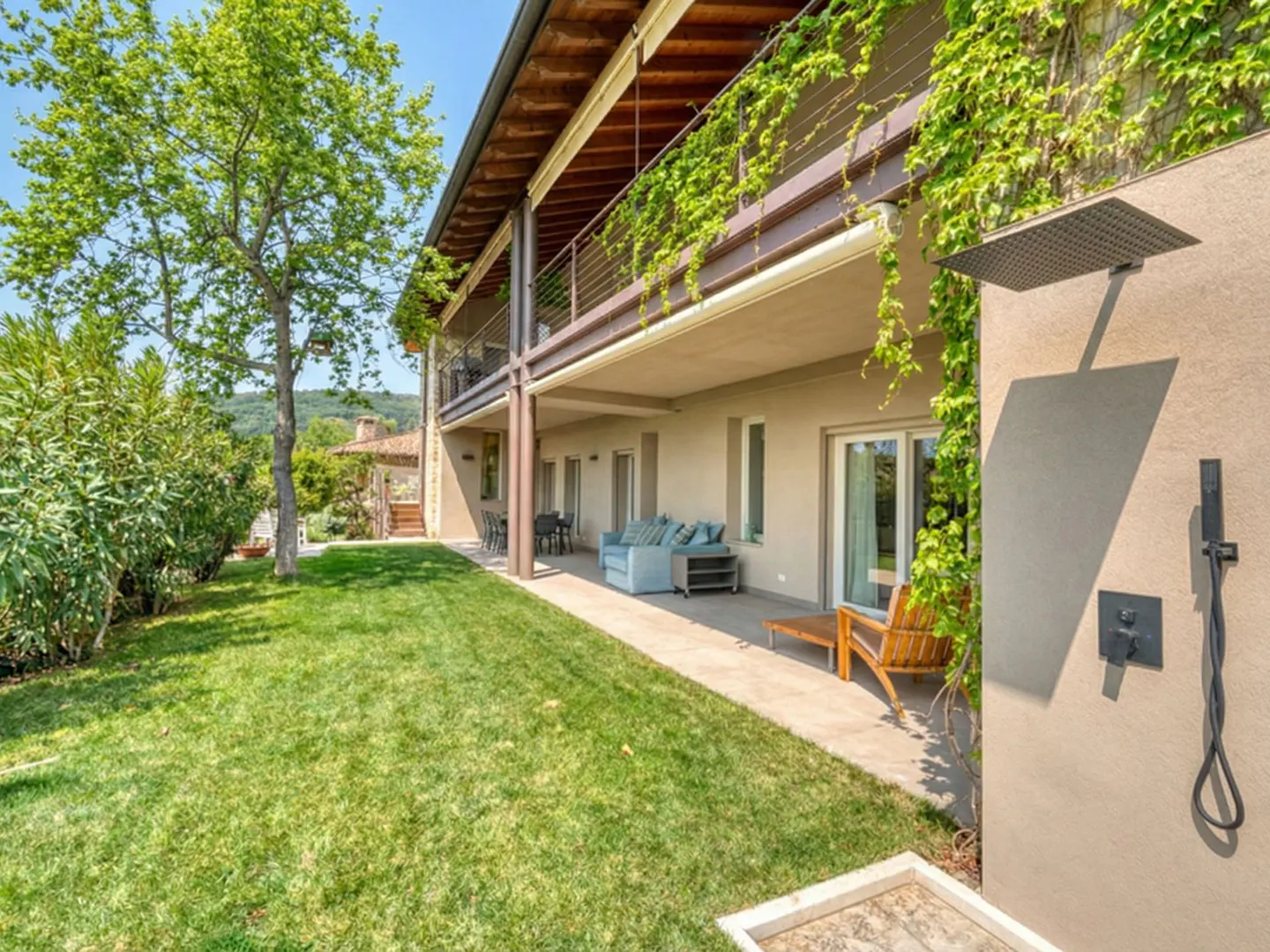 Exterior view of a two-story house with a green lawn, outdoor shower, and patio furniture. Vines climb the building's facade.