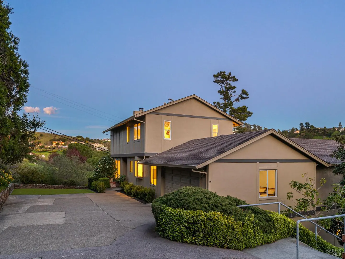Two-story beige house with a gray roof, green bushes, and a concrete driveway at dusk. Lights are on inside.