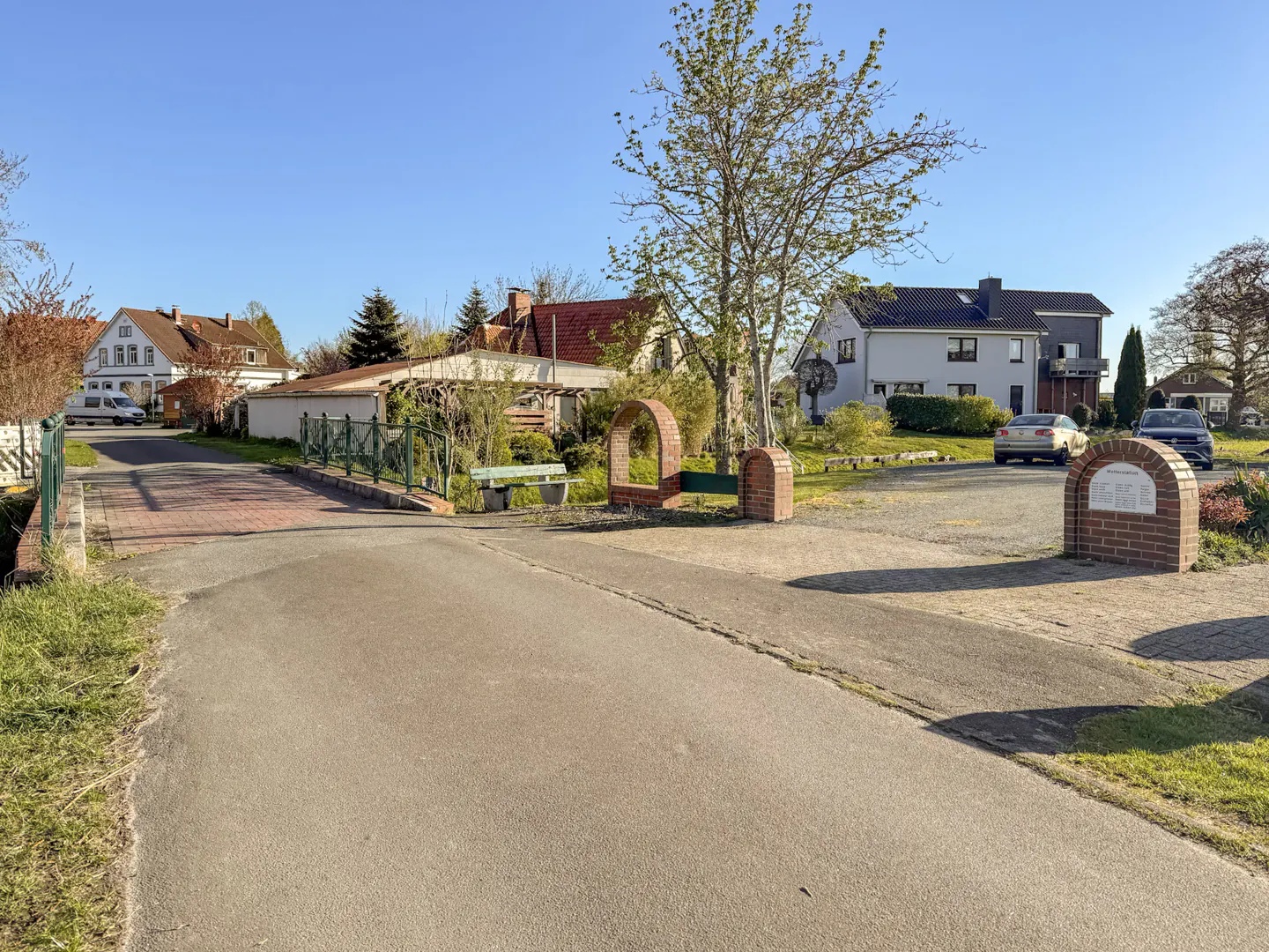 A street view of a quiet neighborhood with houses, trees, and parked cars under a clear blue sky.
