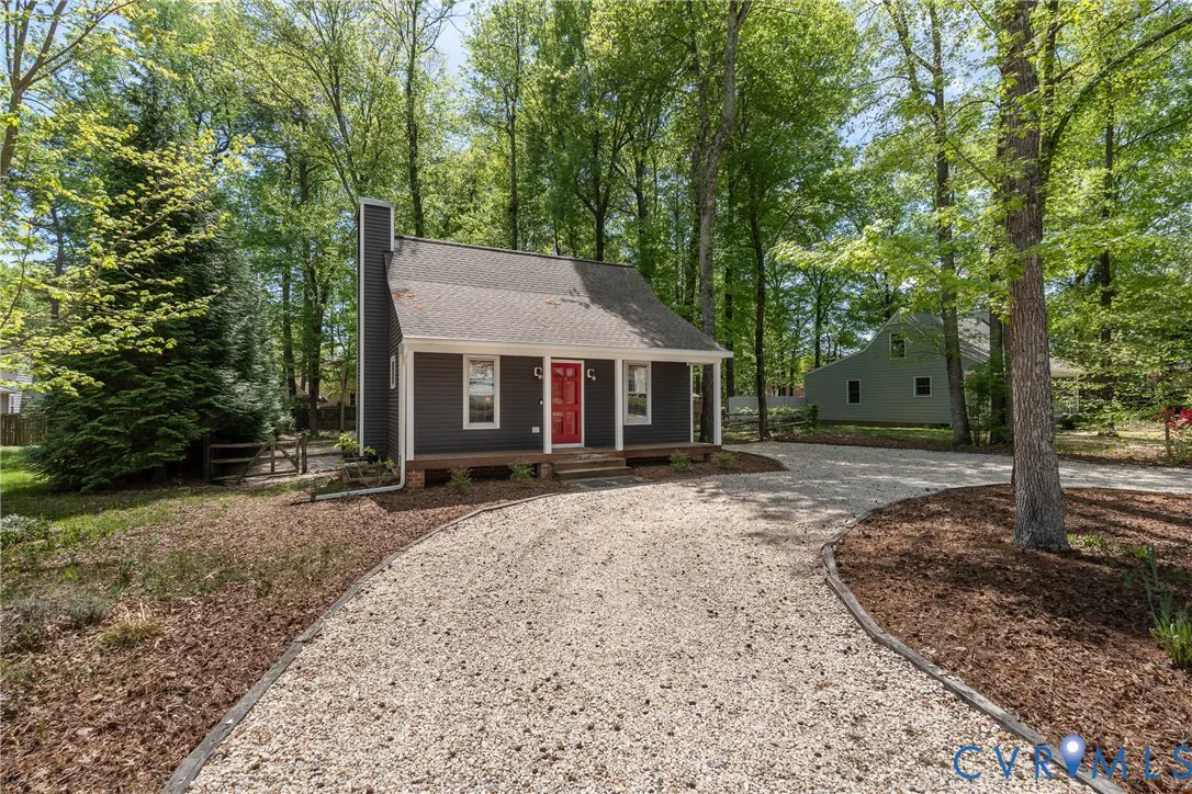 A small, gray house with a red door sits among green trees. A gravel driveway leads to the house.