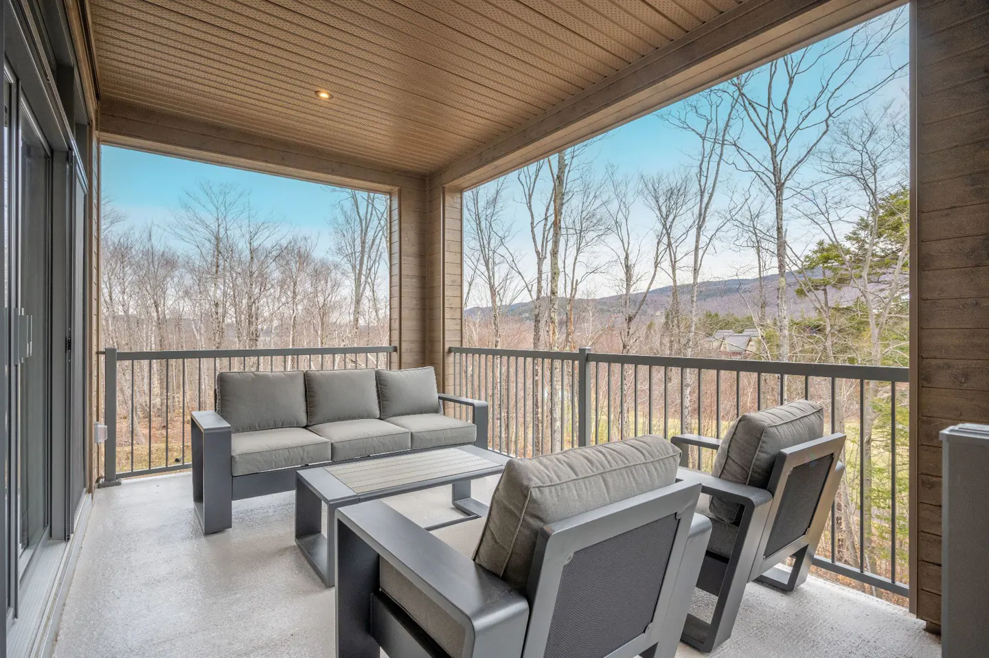 Covered balcony with gray sofa, chairs, and table. Railing overlooks trees and a distant mountain under a blue sky.