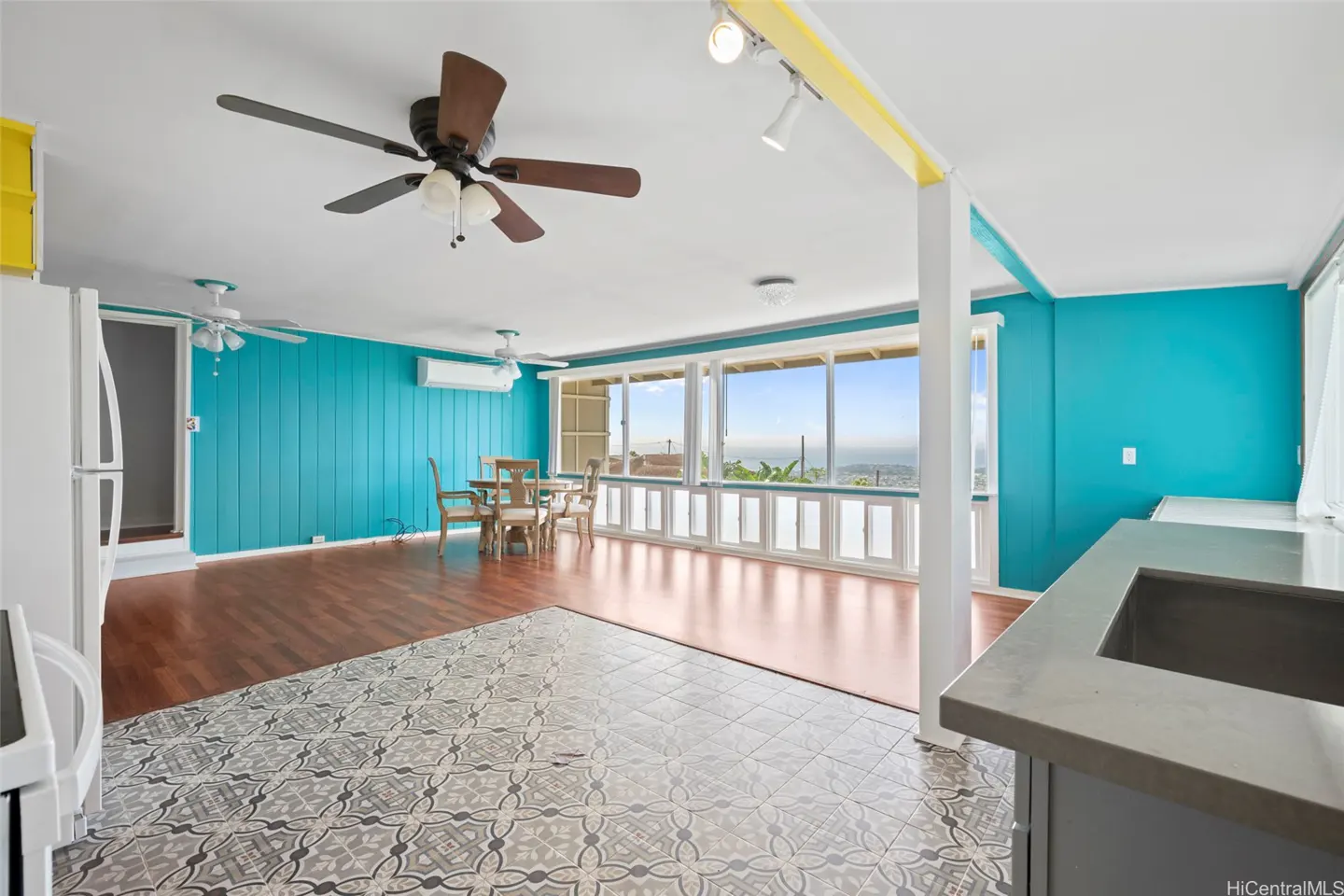 Open-concept living space with turquoise walls, wood floors, and patterned tile. A dining table sits by large windows with an ocean view.