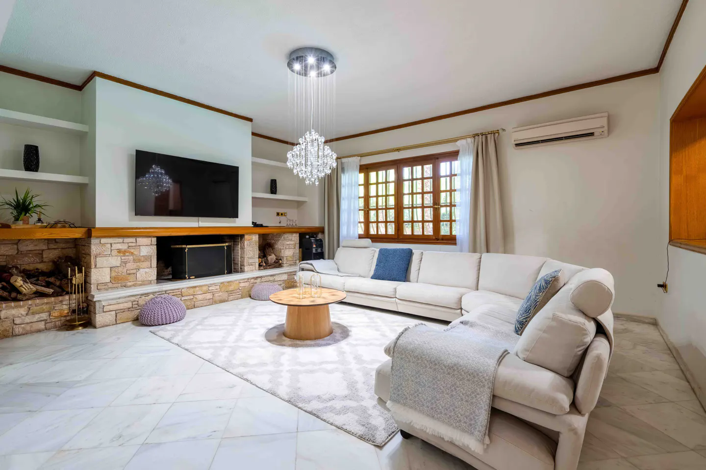 Bright living room with white marble floors, a stone fireplace with a TV above, and a white sectional sofa. A crystal chandelier hangs from the ceiling.