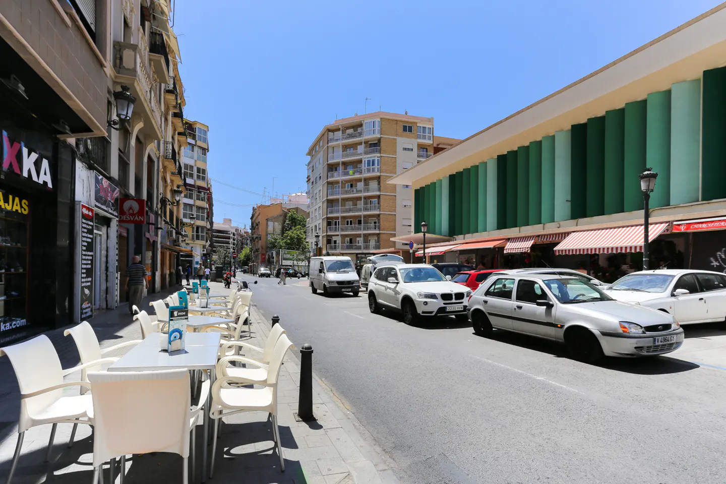 Street view with cars, buildings, and outdoor cafe seating. White tables and chairs line the sidewalk. Cars drive on the street.