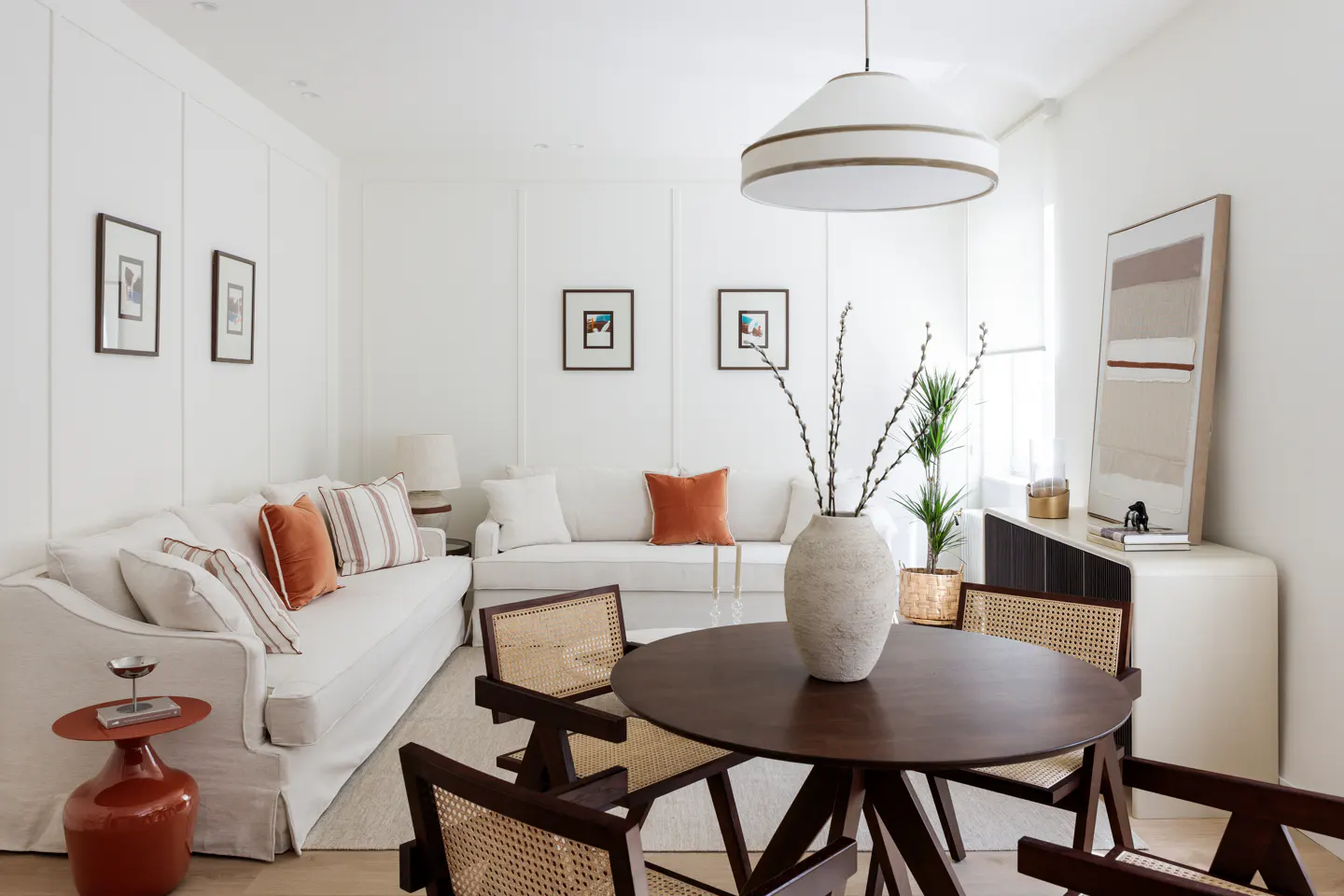 Bright living room with white walls, sofas, and a round dark wood table with four cane chairs. A large white vase sits in the center.