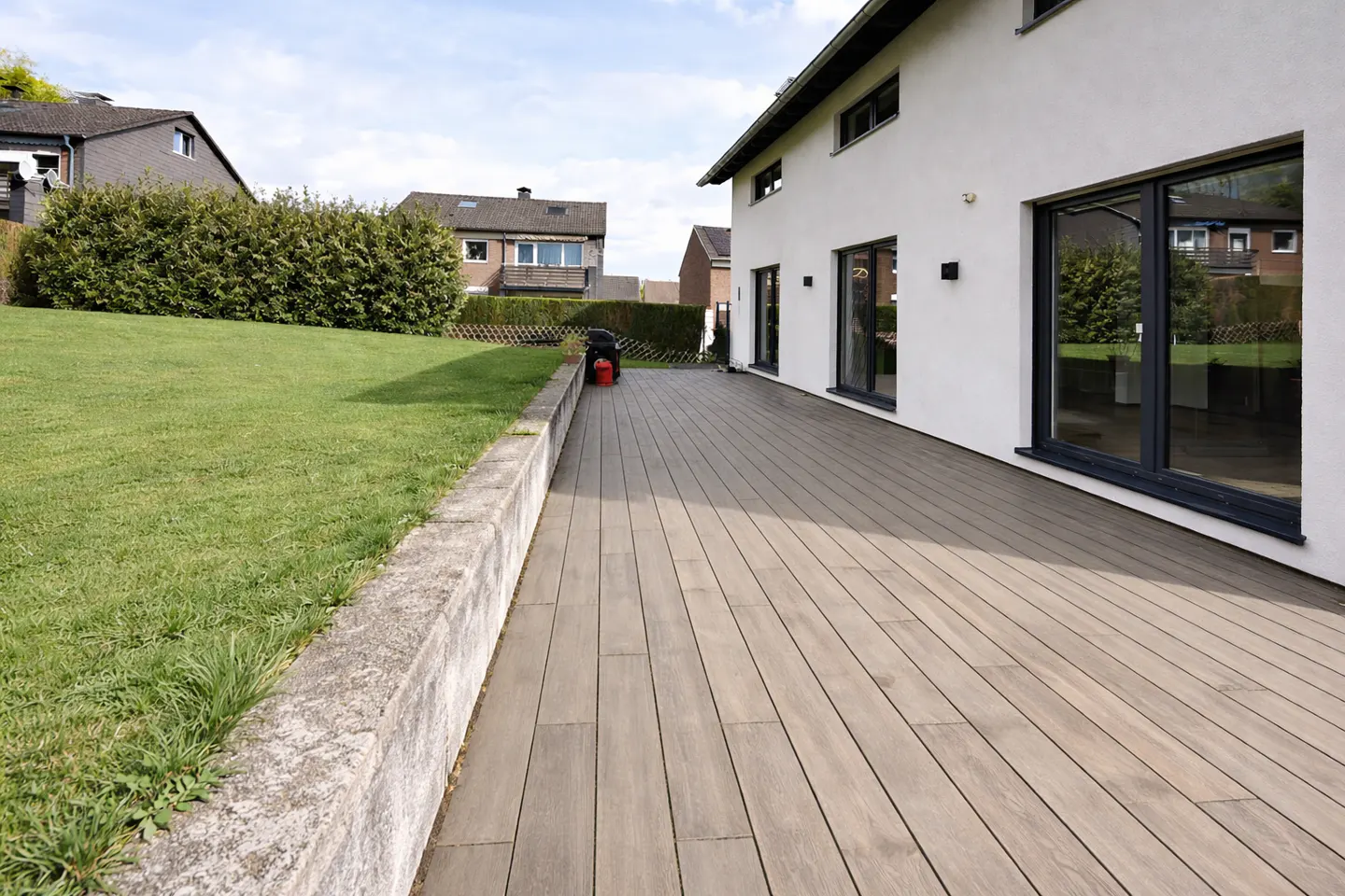 Exterior view of a modern white house with a wooden deck, green lawn, and a retaining wall.