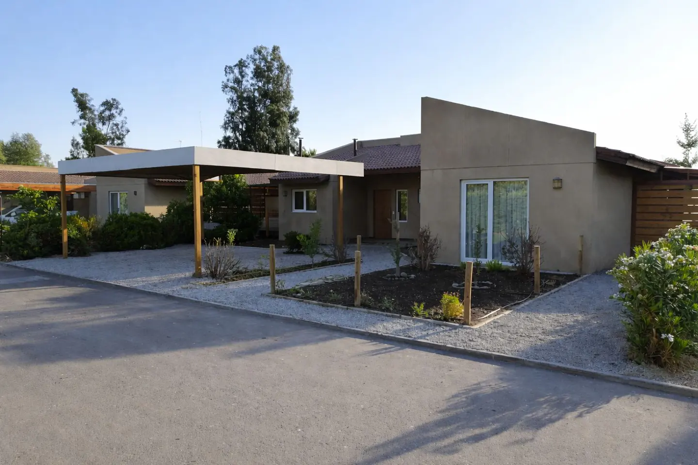 A tan, one-story house with a carport and gravel driveway on a sunny day. The house has a red tile roof and a modern design.