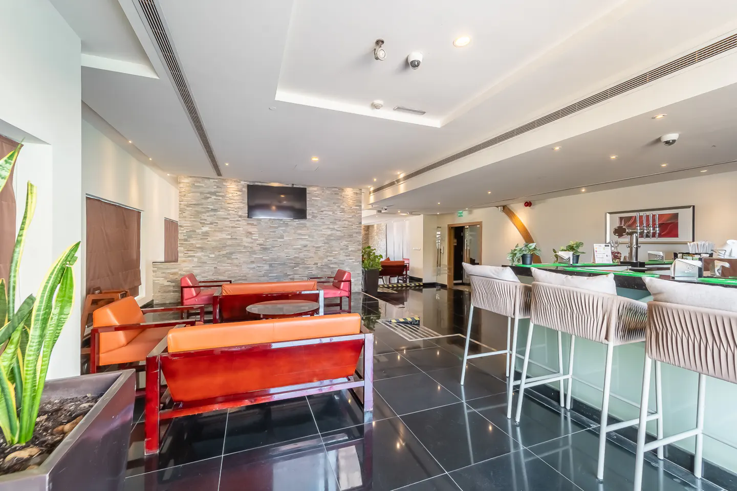 Hotel lobby with orange sofas, bar stools, black tile floor, and a stone accent wall with a TV.