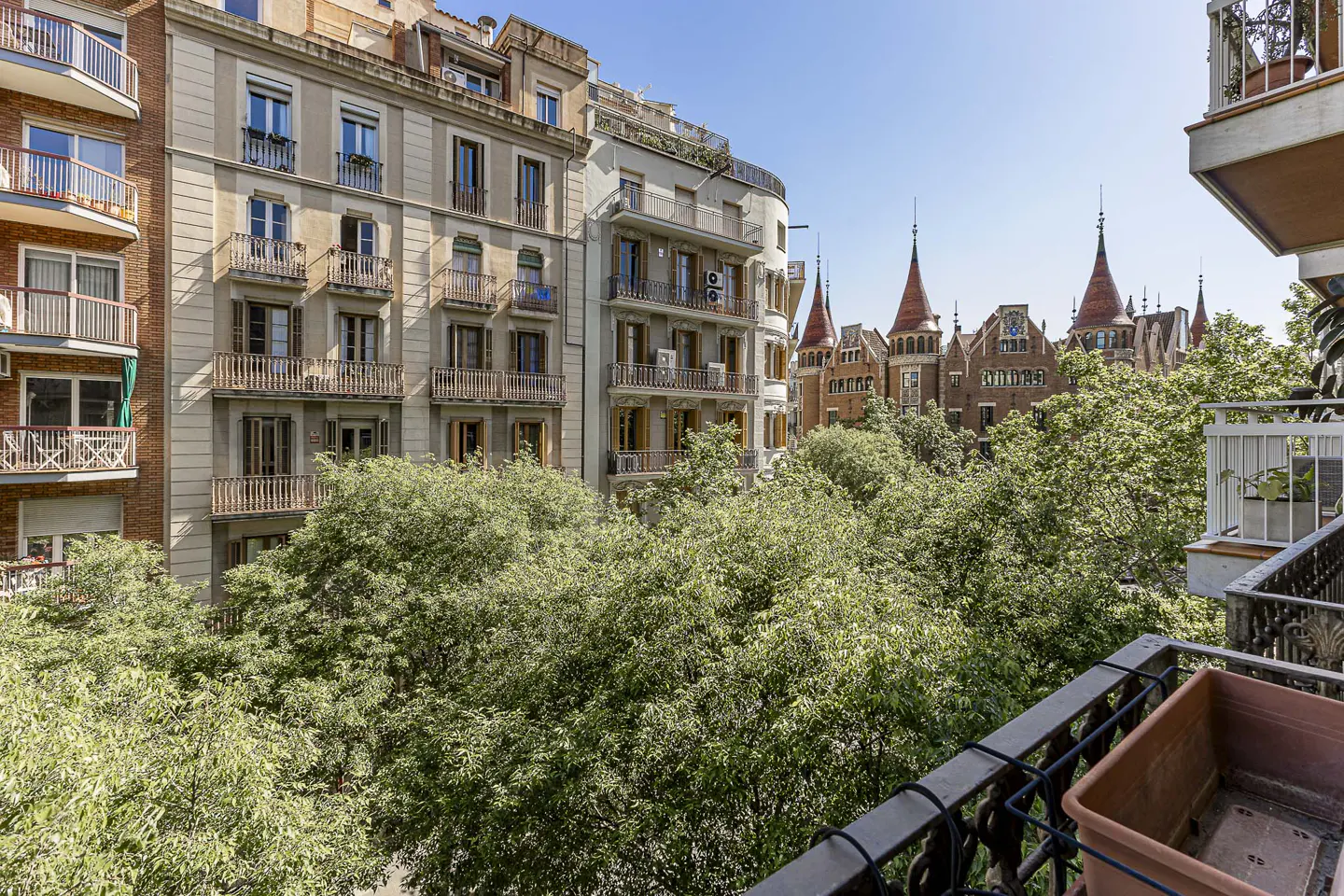 View from a balcony overlooking green trees and buildings in Barcelona, Spain. The Casa de les Punxes is visible in the background.