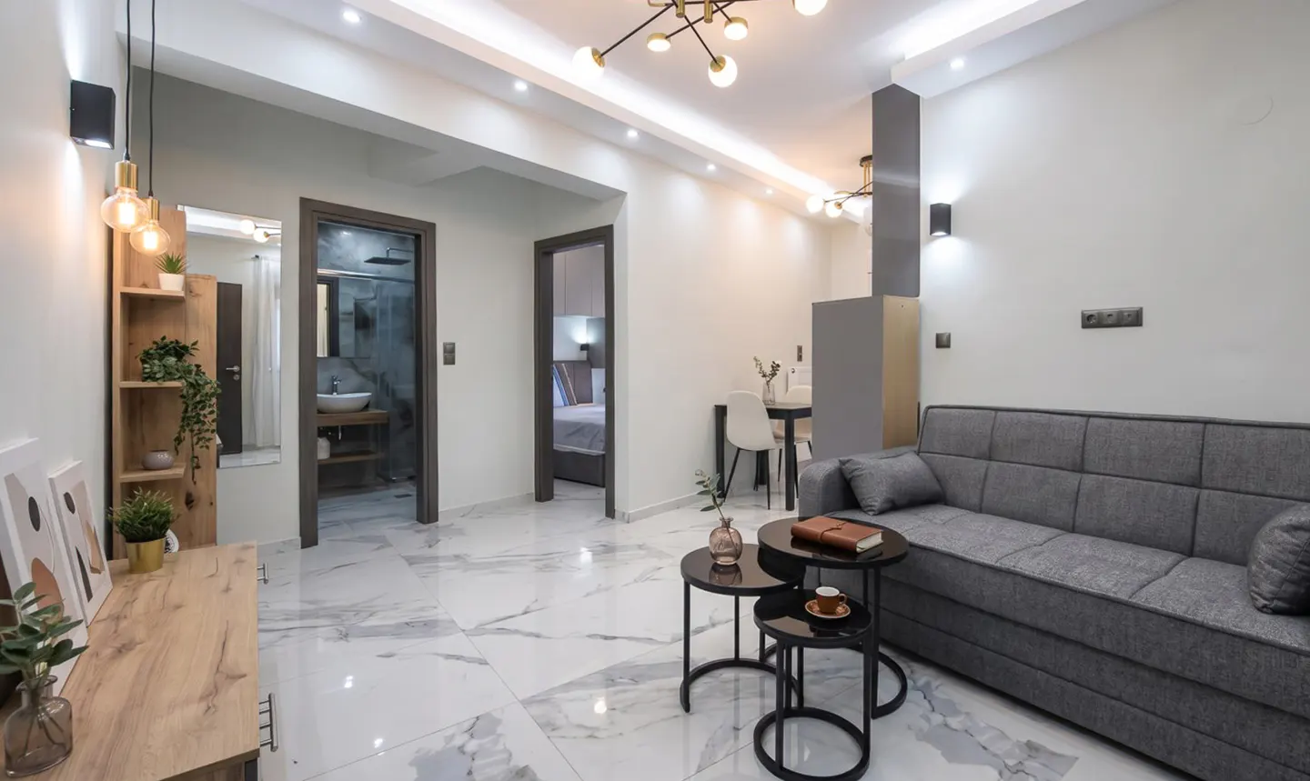 Living room with gray sofa, black nesting tables, and marble floors. Doors lead to a bathroom and bedroom. Modern lighting fixtures.