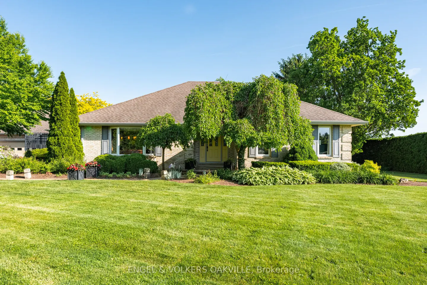 Single-story brick home with a yellow front door, surrounded by lush green trees and a well-manicured lawn under blue sky.