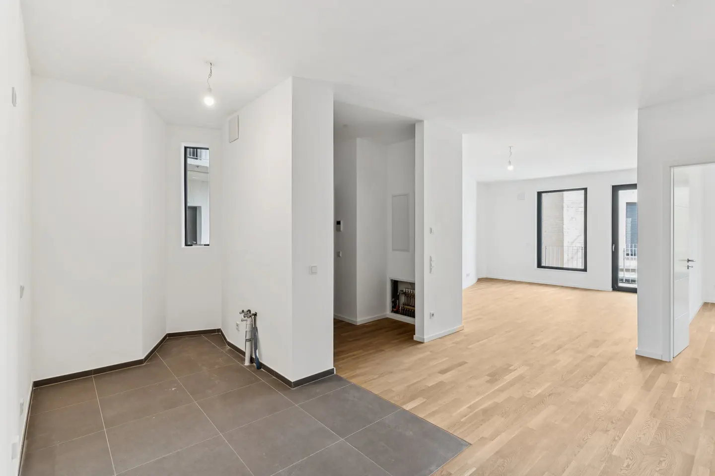 Bright, empty apartment with white walls, light wood floors, and gray tile. A small window and doorway are visible.