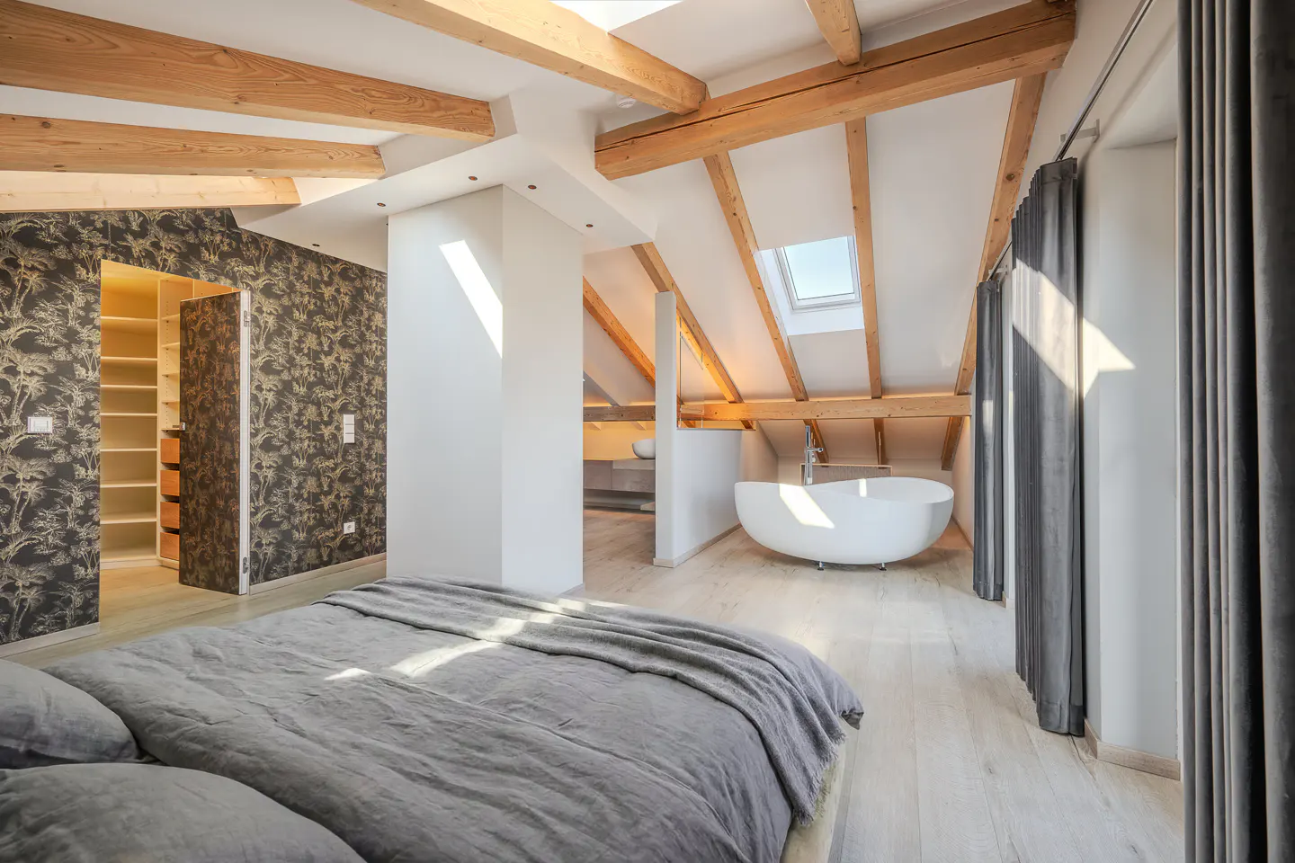 Attic bedroom with exposed wood beams, a gray bed, and a white bathtub. A walk-in closet and gray curtains are also visible.