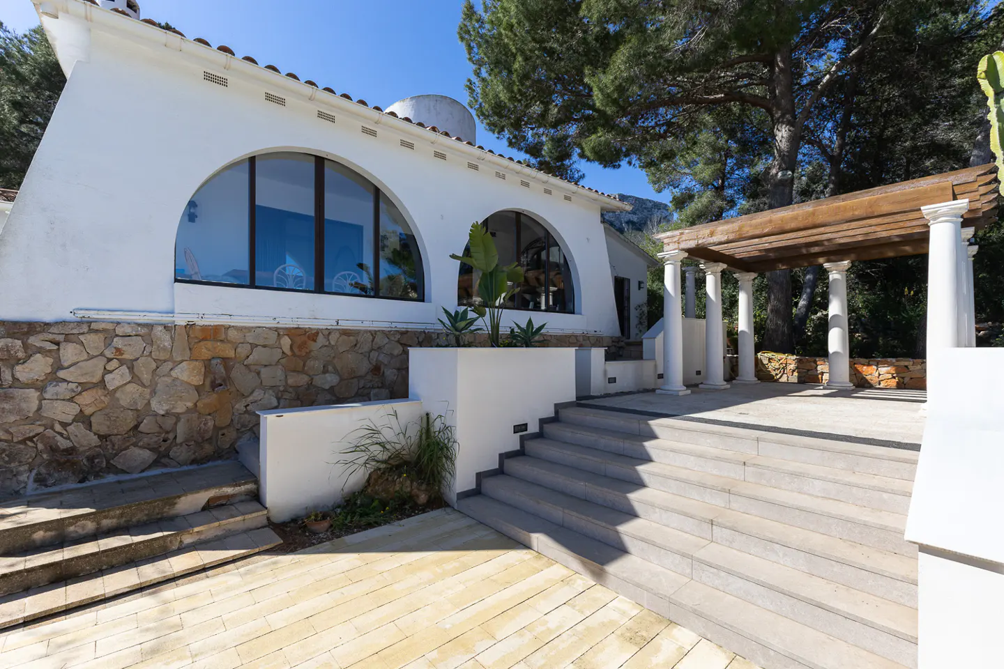 Exterior view of a white house with arched windows, stone accents, and a pergola with white columns. Steps lead to a patio area.