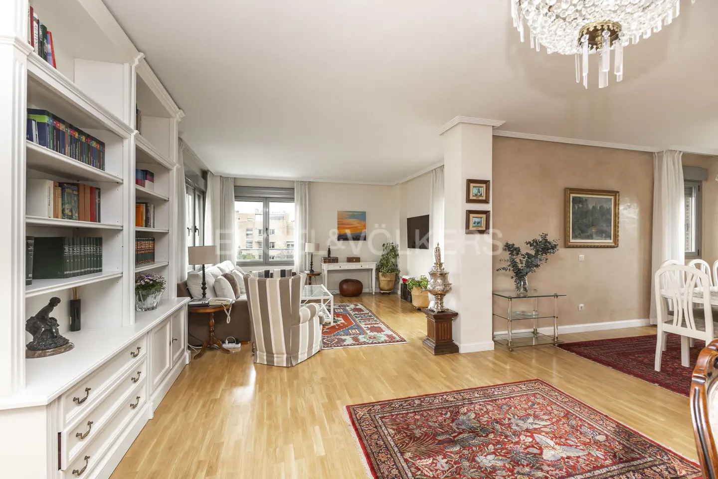 Bright living room with hardwood floors, white built-in bookcase, striped armchair, and red patterned rugs. Chandelier hangs from the ceiling.