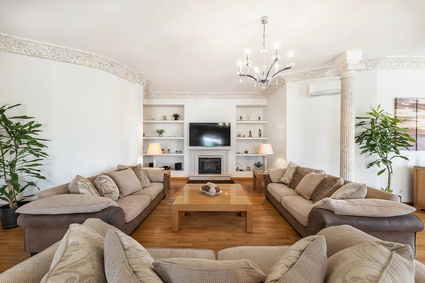 Living room with hardwood floors, white walls, and beige sofas facing a fireplace with a TV above. A chandelier hangs from the ceiling.
