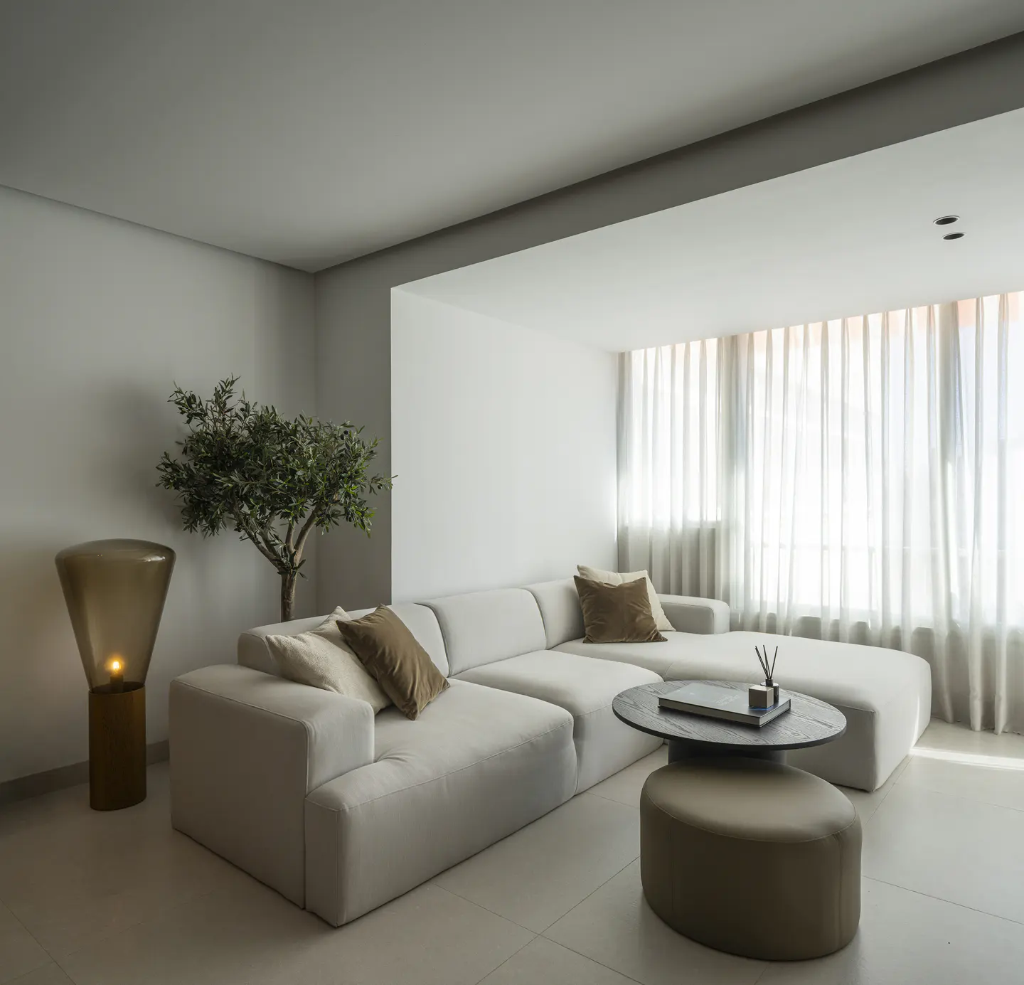 Modern living room with a beige sectional sofa, round coffee table, ottoman, floor lamp, and a small tree by the window.